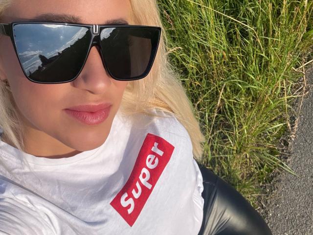 LunaSky-hot with blonde hair, black sunglasses, pink lipstick, white shirt with red "Super" text, black leather pants, and grass in background. Her face is slightly tilted upwards.