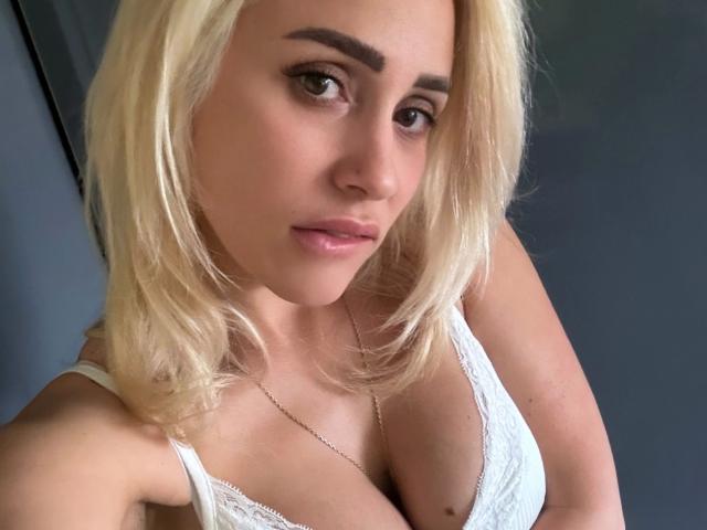 LunaSky-hot with blonde hair, brown eyes, and pink lips, wearing a white lace bra, showing her boobs, and a silver necklace, looking at the camera with a slight smile.