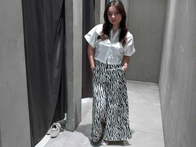AngellMoore with long brown hair, white short-sleeve shirt, and black-and-white zebra-pattern pants, hands in pockets, standing in a gray-walled, dimly-lit room, with white shoes on floor.