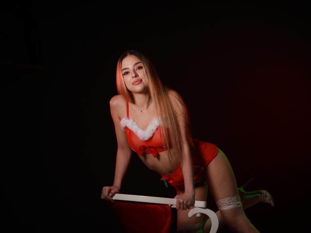 Scarlet Coles, con lunghi capelli biondi, indossa lingerie rossa con bordi in pelliccia bianca e un laccio, appoggiata a una sedia rossa, sorridendo, mostrando il seno e i glutei, in una stanza buia..