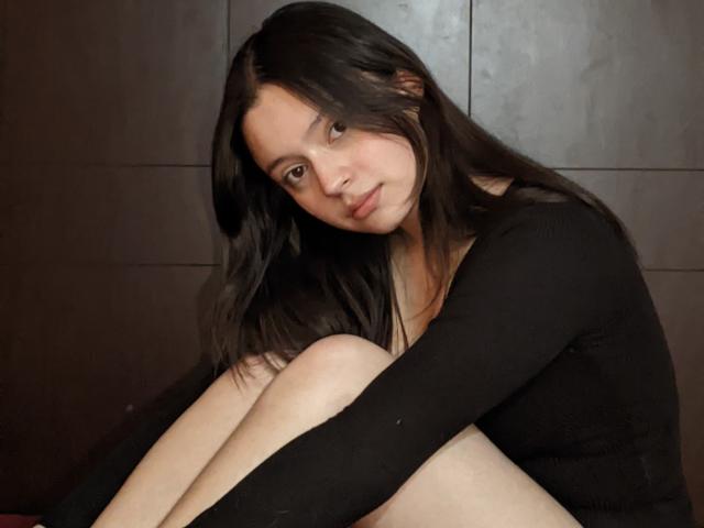 AngellMoore with long black hair, sitting, hugging knees, wearing a black long-sleeve top, looking at camera with a slight smile, brown eyes, fair skin, relaxed pose.