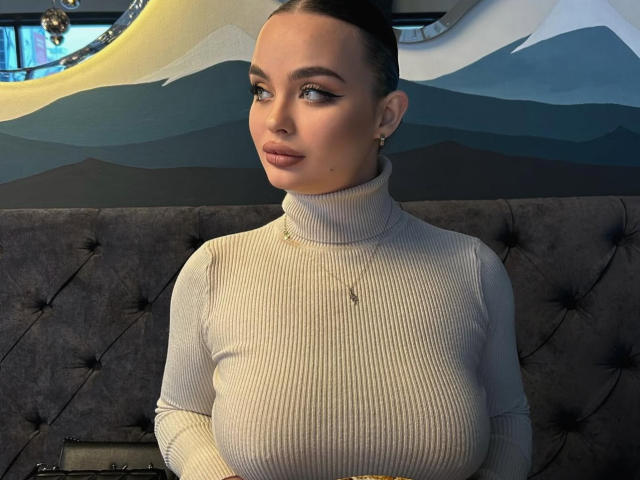 StarStorm with big tits, wearing a ribbed, turtleneck, white sweater, has dark hair in a bun, light makeup, and a small nose ring, sitting on a tufted, gray couch.
