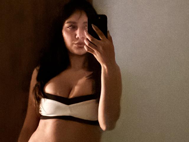DianaFernandezz, avec de longs cheveux noirs, tenant un téléphone, portant un soutien-gorge blanc et noir, montrant ses grandes seins, prenant un selfie devant un miroir, dans un éclairage tamisé..
