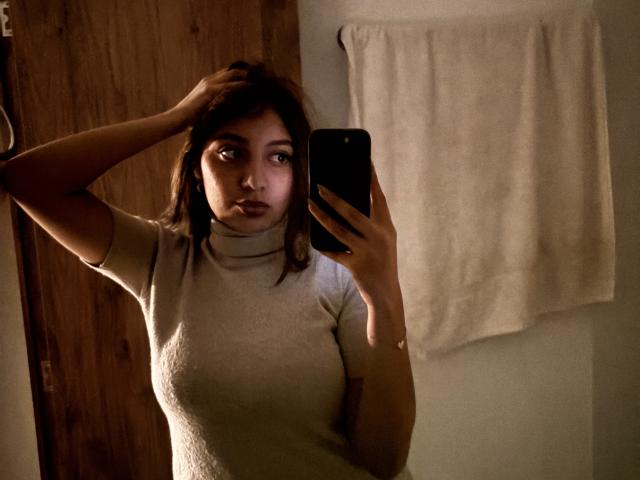 DianaFernandezz with dark hair, wearing a white turtleneck, holding a phone, hand in hair, looking at mirror, medium tits, soft lighting, towel in background, wooden door.