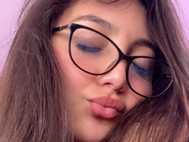 DianaFernandezz with puffy blue eyeshadow, black-rimmed glasses, and slightly parted pink lips, giving a pouty expression with her brown, wavy hair covering part of her face.