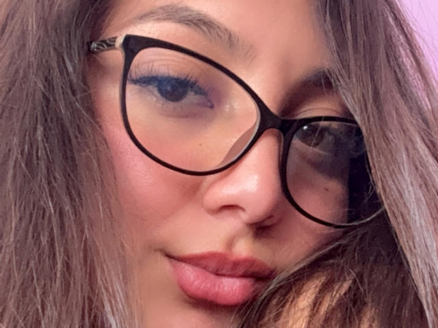 DianaFernandezz's face with black, thick-rimmed glasses, pink lips, and light brown, slightly wavy hair covering one eye, looking slightly to the side with a subtle, confident smile.