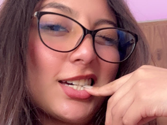 DianaFernandezz con gafas, cabello castaño y lápiz labial rosa, guiñando ligeramente, mordiéndose el dedo cerca de los dientes, mostrando una pequeña sonrisa, con sombra de ojos azul..