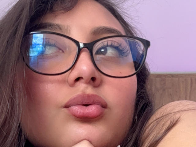 DianaFernandezz with puffy pink lips, dark brown wavy hair, black glasses, and slightly raised eyebrows, looking to the side with blue reflection in her glasses.