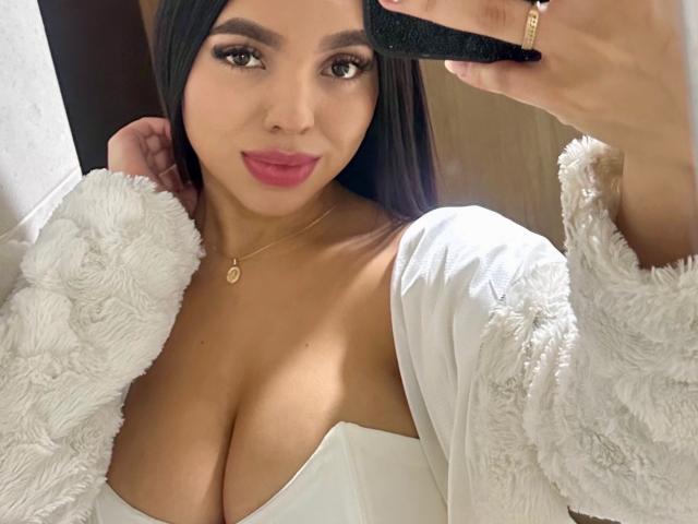 EmilyHotTorres with tan skin, black hair, and red lipstick, wearing a fluffy white cardigan, white dress with cleavage, holding a phone, smiling at the camera.