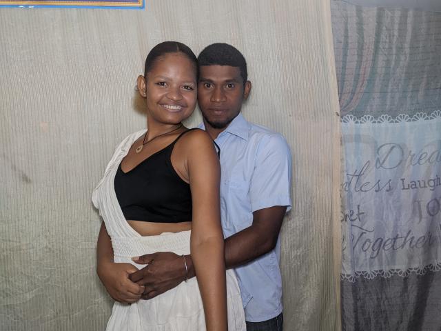 MeriAndMark with small, dark tits, wearing a black sports bra and white drape dress, standing with arms around MeriAndMark's waist, in a light blue shirt.