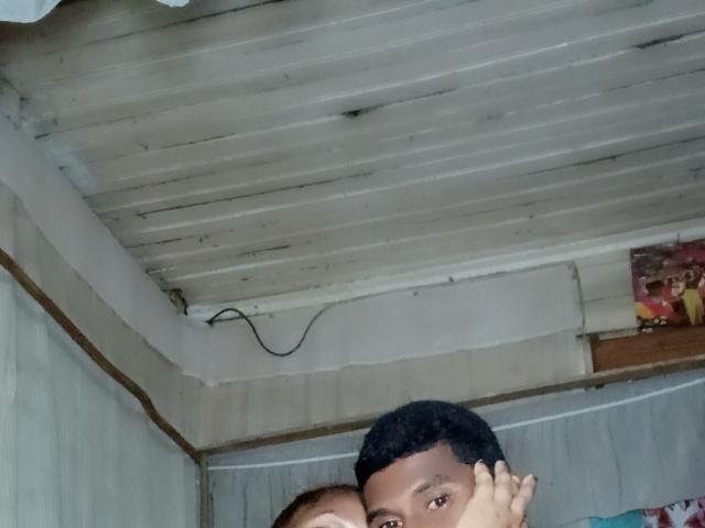 MeriAndMark with short black hair, holding a small child's face, visible only from the nose up, in a dimly lit, wooden ceiling room.