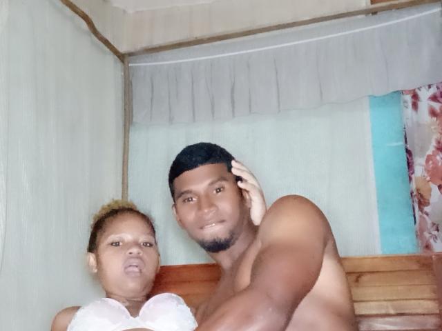 MeriAndMark with dark skin and short black hair, shirtless, next to a person with light brown skin, curly hair, and white shirt, in a wooden bed.