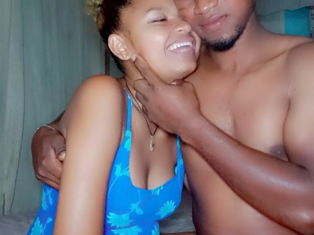 MeriAndMark with tan skin, smiling, wearing a blue floral tank top, and Mark with dark skin, touching her neck, both topless, in a cozy, intimate setting.