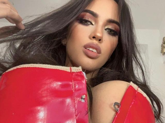 AlexaPaulfer with long dark hair, red shiny leather top, and heavy makeup, showing off her big, shiny tits, holding her hair, looking sexy with parted lips.