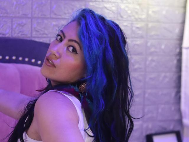 Ailich with blue and black wavy hair, looking over shoulder, nose ring, eyeliner, white top, purple-pink light, and gold earrings, showing her ass slightly.