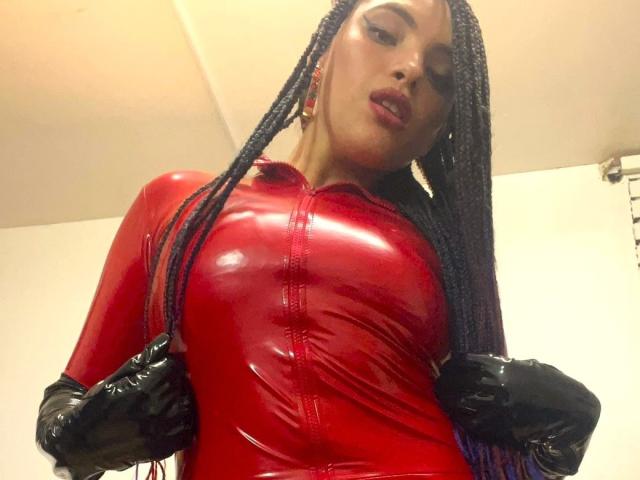 ElizaLovely with long braids, wearing a shiny red latex dress, black latex gloves, and looking down at the camera, showing off her big tits and confident expression.