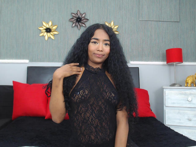 CrithyMarks with long black curly hair, wearing a black lace halter top, showing her small black tits, smiling, standing in a bedroom with red pillows and a red lamp.
