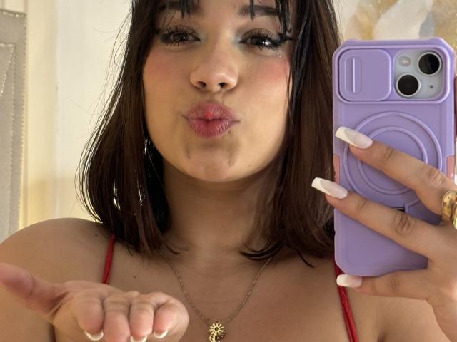 MillyJohnson with pouty lips, brown shoulder-length hair, and white nails, holding a purple phone, red strap visible, pointing with index finger, gold necklace with flower pendant.