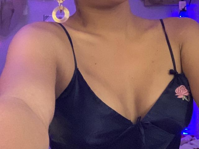 ErikaTS with tan skin, wearing a black satin camisole with pink rose embroidery, showing her boobs, wearing large gold hoop earrings, in a dimly lit room.