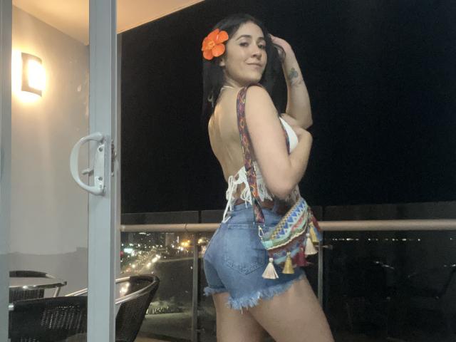 NicoColeman with black hair, orange flower in hair, backless top, fringed denim shorts, colorful bag, standing on balcony at night, smiling, showing her ass, tattoos on right arm.