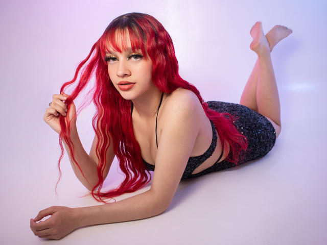 Ada Ferreira mit roten, gewellten Haaren, liegt auf dem Bauch, trägt ein schwarzes, glitzerndes Kleid, das ihre Brüste und ihren Hintern zeigt, die Beine sind angehoben, sie hält sich die Haare, weißer Hintergrund, niedlich..