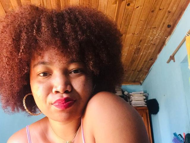 Orabelle with big brown afro, pink lipstick, hoop earrings, and dark skin, looking at camera, shoulder up, wooden ceiling, and blue wall in background. Her tits are small.
