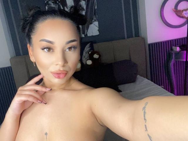 MadyLux, senza top, con seni di medie dimensioni, capelli scuri raccolti in chignon, occhi marroni, labbra rosa, tatuaggio sul braccio destro, mano sul petto, in camera da letto, arredamento viola e grigio, che guarda verso la fotocamera..