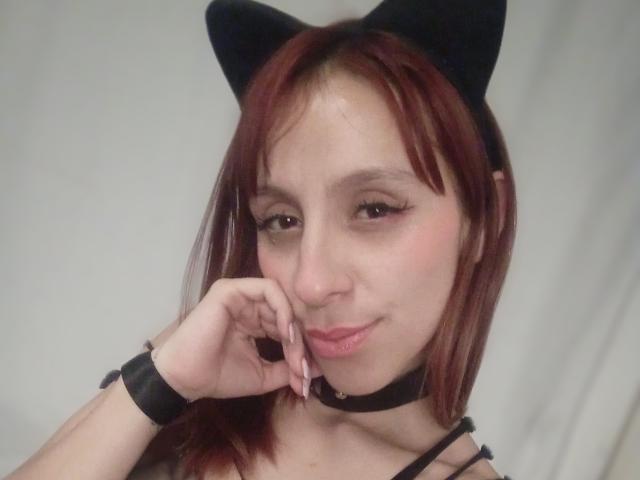 DanielaKeiko, avec des cheveux roux, des oreilles de chat noires, un collier noir et un bracelet, appuyant sa joue sur sa main, souriant avec du rouge à lèvres rose, et ayant une allure à la fois mignonne et séduisante..