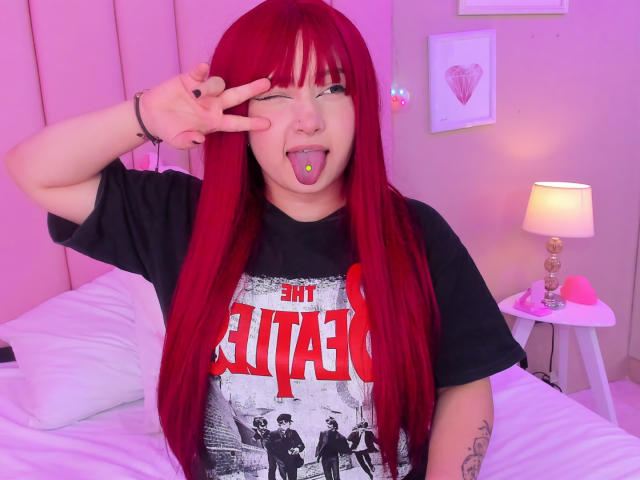 TracieLowe with bright red hair, winking, sticking out her tongue with a green piercing, wearing a black "Eagles of Death" shirt, making a peace sign.