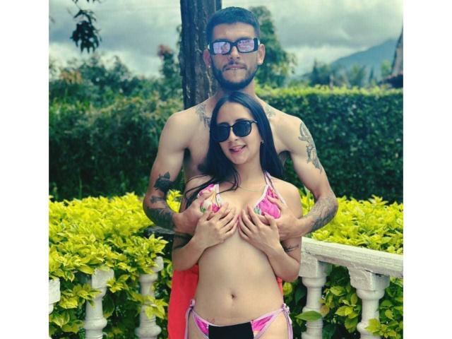GrettaAndDannie in pink bikini, Dannie shirtless with tattoos, standing behind, hands on Gretta's tits, both wearing sunglasses, green hedge and white railing in background.