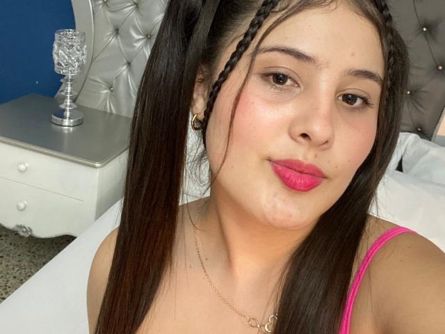 Kathy Stone, com lábios vermelhos e inchados, rabos de cavalo pretos, vestindo um top rosa de alças finas, um colar de ouro e brincos pretos, fazendo uma careta. Seus seios estão ligeiramente visíveis..