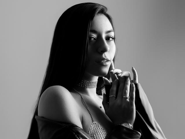 La partie supérieure de la poitrine et le cou de CanelleGarces sont visibles, avec un collier scintillant et une fine chaîne, ses longs cheveux noirs et un maquillage discret. Les seins sont partiellement visibles. Noir et blanc..