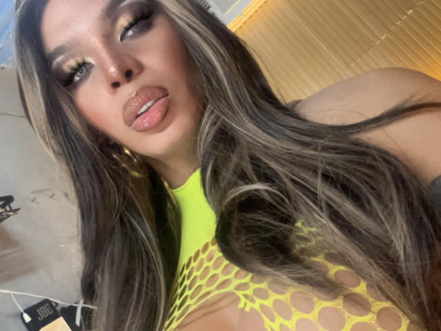 BigCockDominantX with long brown hair, yellow mesh top showing big tits, pouty lips, and gold earrings, holding hair, looking at camera.