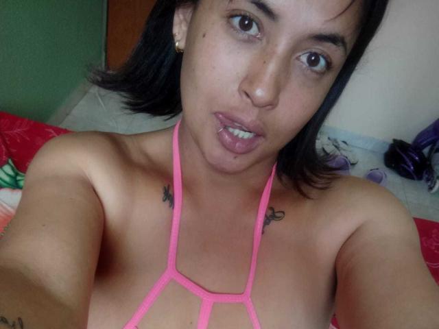 MirandaDreams with short black hair, wearing a pink strappy top, showing small tattoos on her shoulders, slightly parted lips, and a subtle facial scar. Her tits are visible.