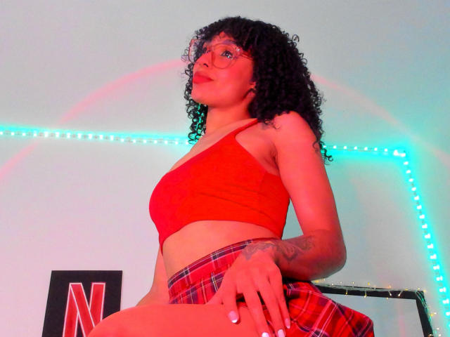 NickiBloom with curly black hair, red glasses, red crop top, plaid skirt, and tattoo on right arm, looking up, red and blue lights, standing, confident, sexy.