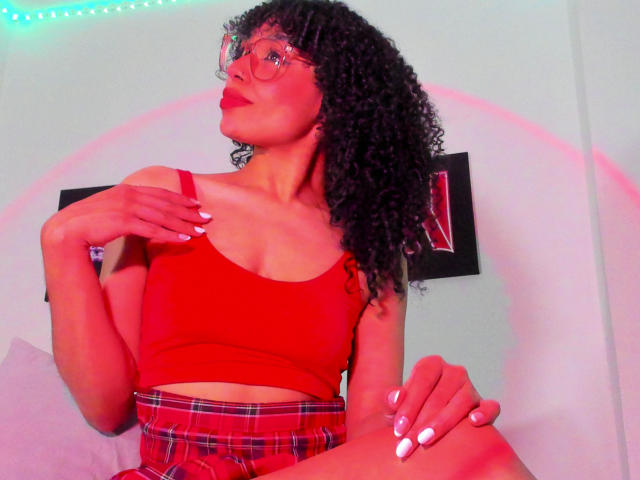 NickiBloom with curly black hair, red glasses, red crop top, and red plaid skirt, showing off her tits and white nails, sitting with a hand on her knee.