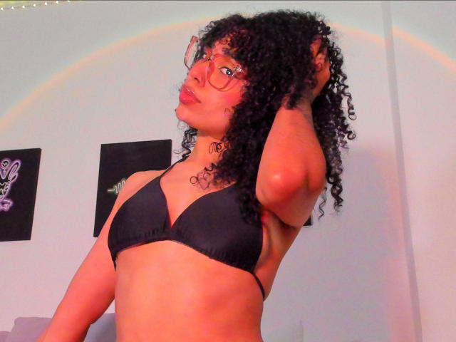 NickiBloom with curly black hair, glasses, and red lipstick, wearing a black bra, hands in hair, rainbow light on pale skin, visible tattoo on neck.