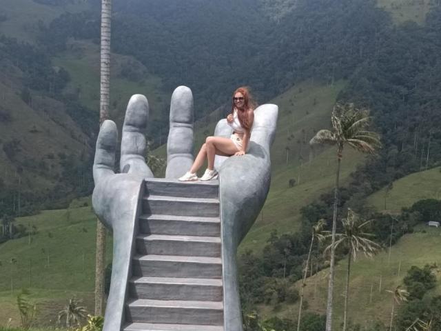 MiaPoe with red hair, sunglasses, white top, and shorts, sitting on a giant gray hand's stairs, surrounded by green hills and palm trees.
