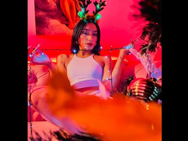 SalomeMille with antlers, wearing a white crop top, sitting with legs spread, holding Christmas lights, red and pink neon lighting, Christmas ball in foreground, looking seductive.