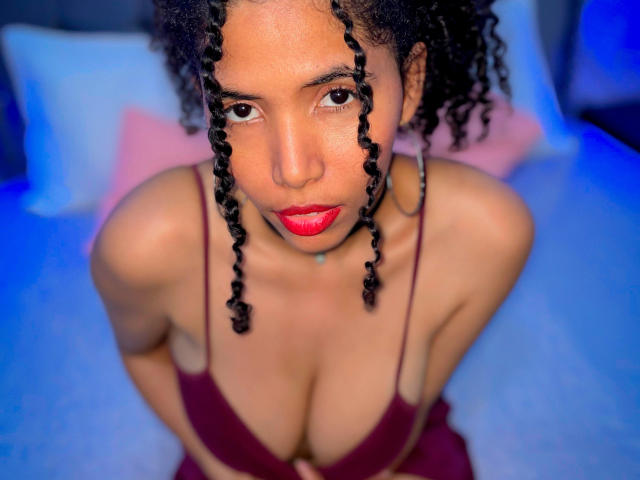 ZoeJonees with curly black braids, red lipstick, and a maroon bra, looking up with her tits prominently displayed, sitting on a bed with blue and pink pillows.
