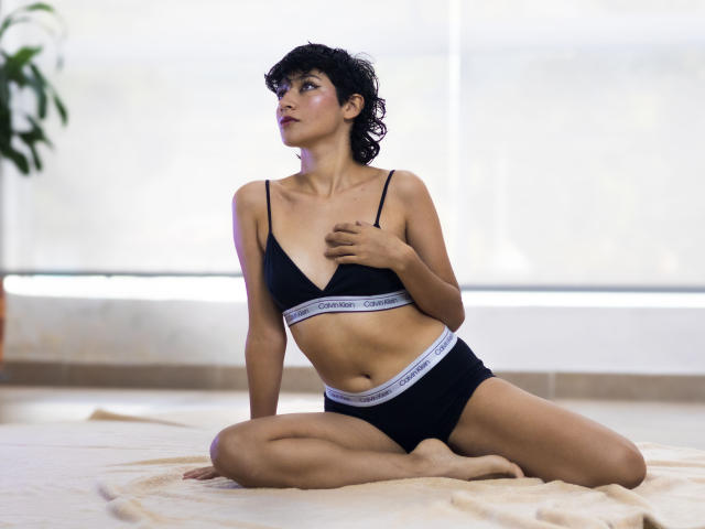 SweetAmbers with short black hair, wearing black Calvin Klein bra and underwear, sits on a beige towel, hand on chest, looking to the side, legs crossed, soft light.