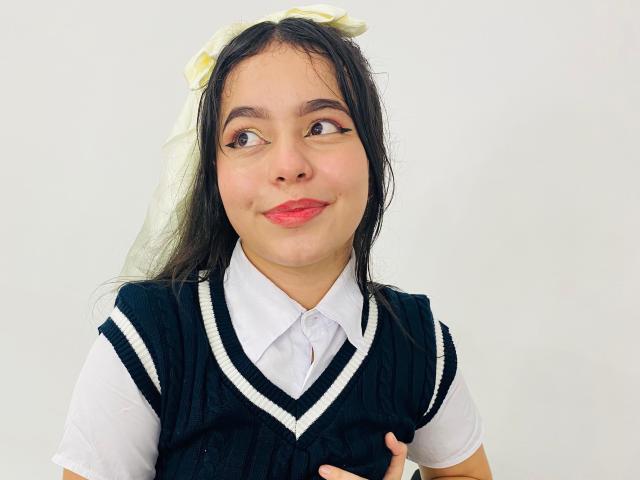 LissyCarter with long black hair, white dress, and black sleeveless top, holding her tits, smiling with red lipstick, and a white flower in her hair, on a white background.