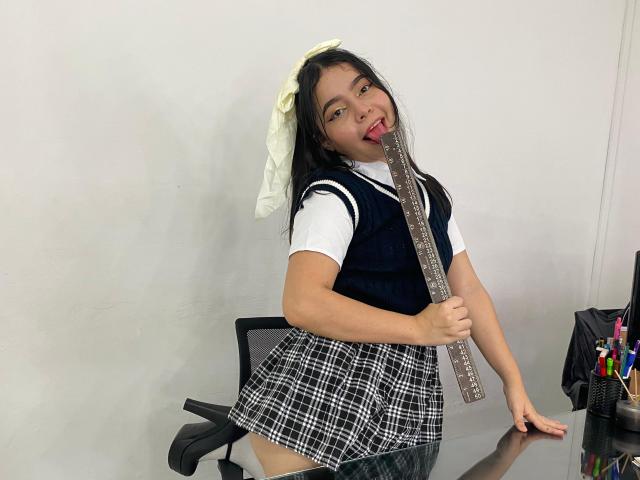 LissyCarter with dark hair, white bow, school uniform, holding ruler in mouth, plaid skirt, black vest, white shirt, standing by desk, smiling, showing teeth, looking cute.