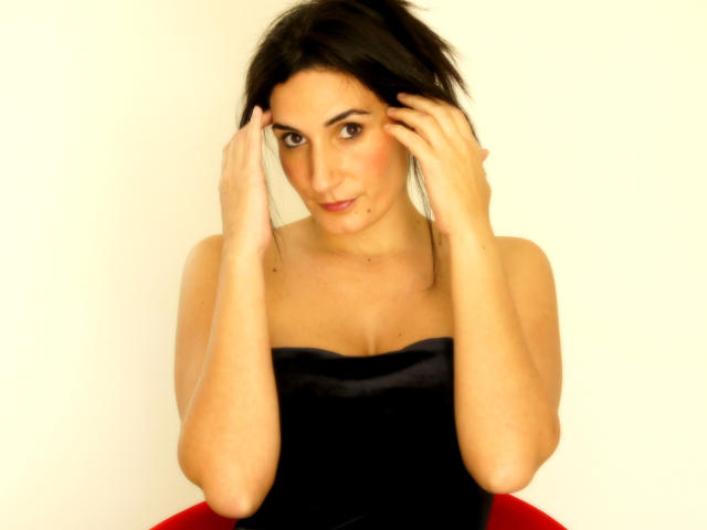 GoddessDolly con capelli neri, pelle chiara e seno piccolo, indossa un top nero senza spalline, mani tra i capelli, leggero sorriso, seduta su una sedia rossa, illuminazione soffusa, guarda la fotocamera..