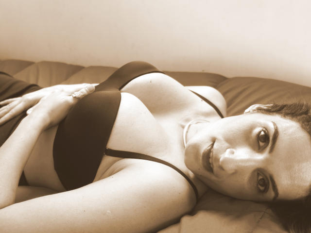 GoddessDolly liegt auf einem Bett, trägt einen schwarzen BH, zeigt ihre Brüste, blickt mit einem leichten Lächeln in die Kamera, die rechte Hand liegt auf ihrem Bauch, sepiafarben..