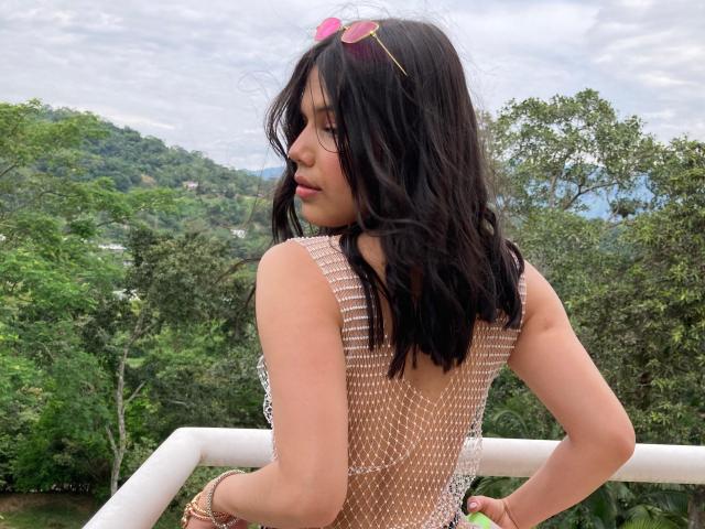 AmyXHottie with wavy black hair, pink sunglasses on head, wearing a see-through mesh top, showing her back and small ass, standing on a white balcony.