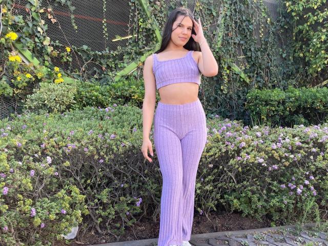 AmyXHottie con una camiseta corta a rayas moradas y pantalones de cintura alta, de pie, con la mano en el pelo, mirando a la cámara, pechos medianos, delgada, piel morena, pelo largo y negro, fondo de hojas verdes..