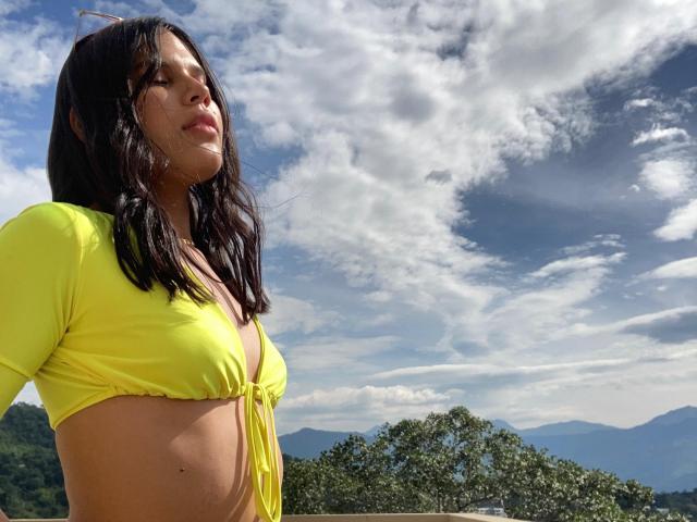 AmyXHottie with tan skin and dark wavy hair, wearing a yellow bikini top, looking up at a cloudy blue sky, mountains in the background, and green trees below.