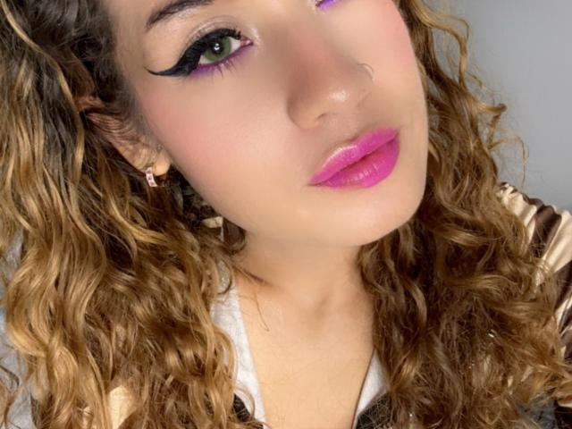 AkiraZanoli has curly brown hair, green eyes with black eyeliner, purple eyelid, pink lipstick, and a nose ring. She's wearing a white shirt with a brown pattern.