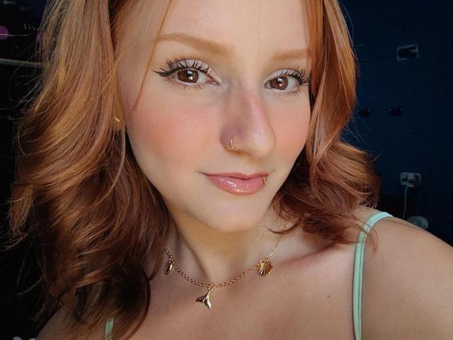 RuivaK has red hair, brown eyes, and is wearing a nose ring and lip piercing. She's sporting a green tank top and a gold necklace with leaf charms.